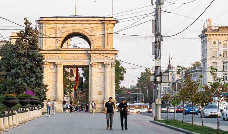 Arch of Triumph, Chisinau, in Republic of Moldova.6 persons visible walking from and towards the arch during daytime. some other buildings visible in the distance, trees and flowers on the left side