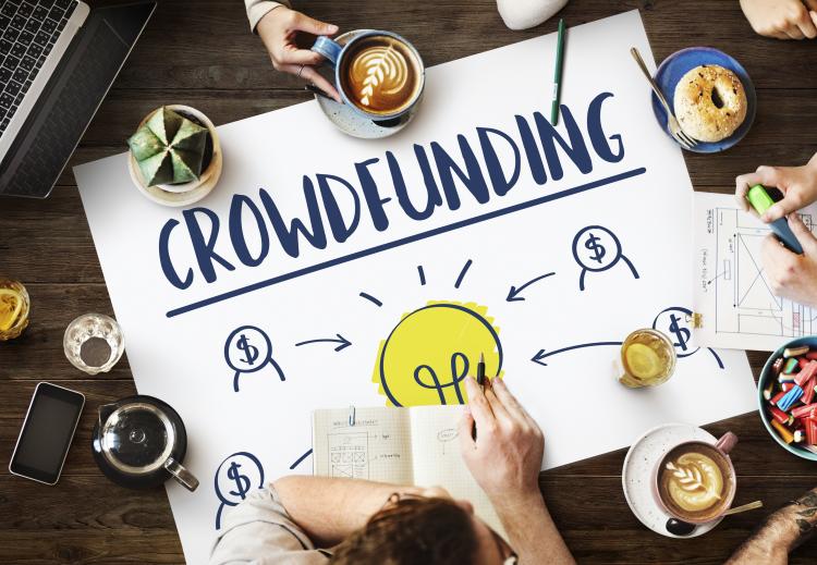 People sitting at a big table, only their hands are visible. in the middle of the table there is a big flipchart where crowdfunding is written, there is a yellow light bulb below. 