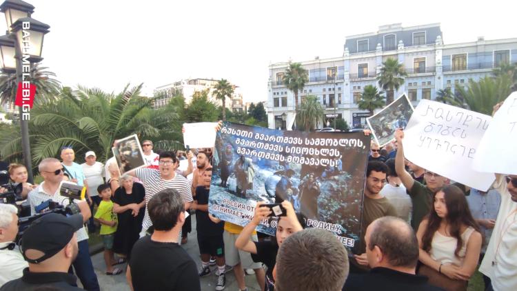 Numerous protestors holding Georgian and Ukranian posters, monumental, light blue building in the background and palm trees on the left side