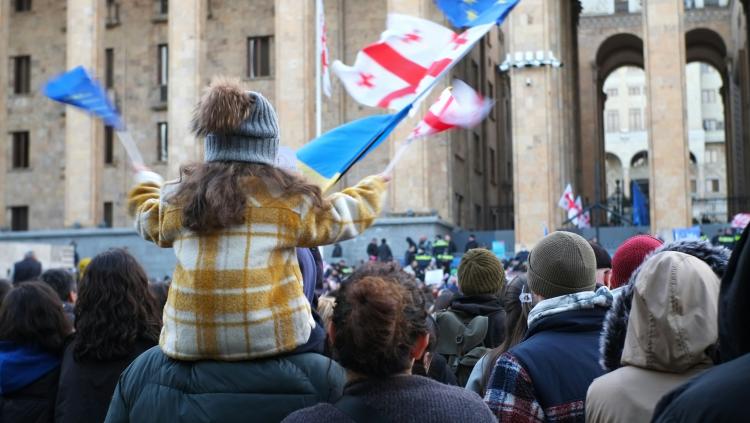 Girl sits on the shoulders of her parent at a protest, surrounded by the flags of Georgia and the European Union.